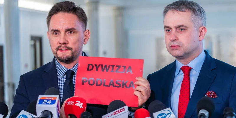 Na zdjęciu dwóch mężczyzn stoi przed mikrofonami, trzymając czerwony plakat z napisem "DYWIZJA PODLASKA".