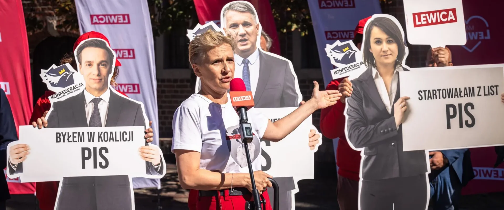Kobieta przemawia do mikrofonu podczas wiecu, otoczona kartonowymi wycinankami polityków oraz plakatami z logotypami Lewicy, Konfederacji i PiS.