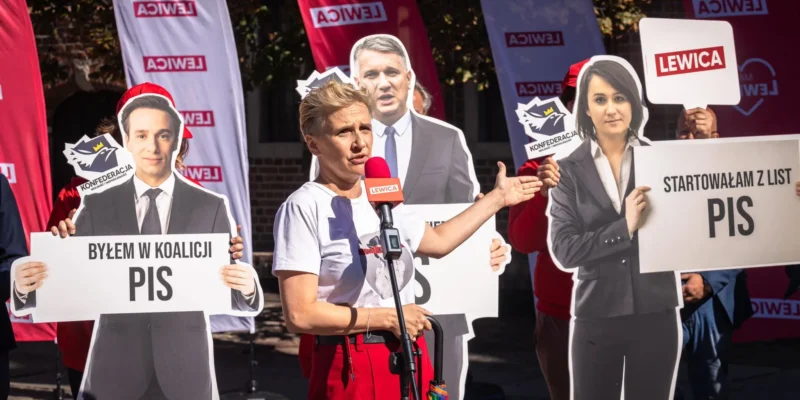 Kobieta przemawia do mikrofonu podczas wiecu, otoczona kartonowymi wycinankami polityków oraz plakatami z logotypami Lewicy, Konfederacji i PiS.