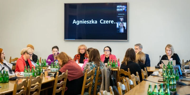Zdjęcie przedstawia zebranie w sali, podczas którego uczestnicy dyskutują, a na dużym ekranie widoczny jest tekst „Agnieszka Czere...”.