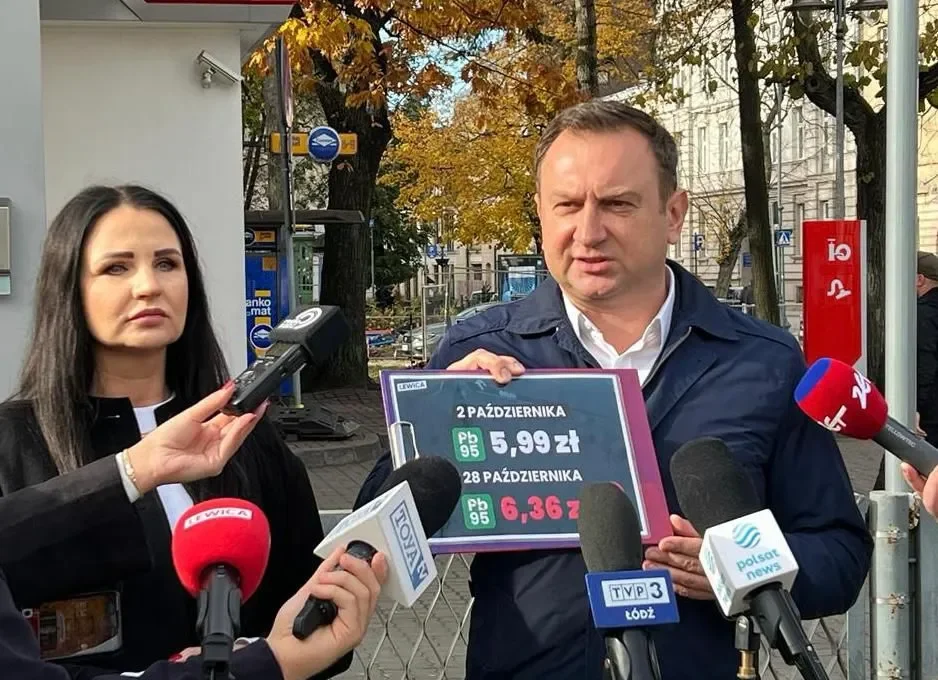 Mężczyzna trzyma tablicę z cenami paliw (5,99 zł i 6,36 zł) podczas konferencji prasowej, otoczony reporterami z mikrofonami przed stacją benzynową.