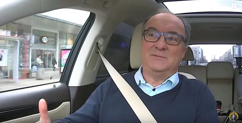 Na zdjęciu widać mężczyznę siedzącego w samochodzie z wyraźnym uśmiechem, który gestykuluje podczas rozmowy.