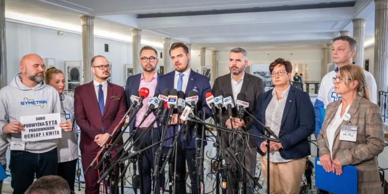 Na zdjęciu grupa osób stojąca przed mikrofonami na konferencji prasowej, z jednym mężczyzną mówiącym do mediów, a inni trzymającymi plakaty.