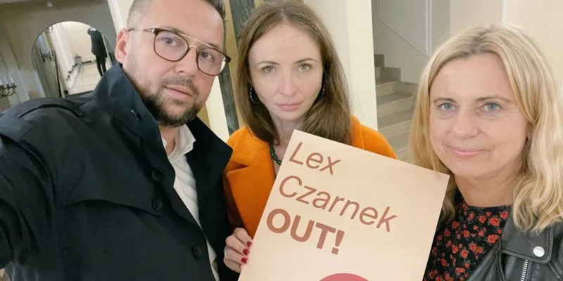 Na zdjęciu trzech ludzi trzyma banner z hasłem "Lex Czarnek OUT!" w neutralnym wnętrzu.