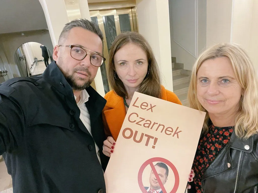 Na zdjęciu trzech ludzi trzyma banner z hasłem "Lex Czarnek OUT!" w neutralnym wnętrzu.