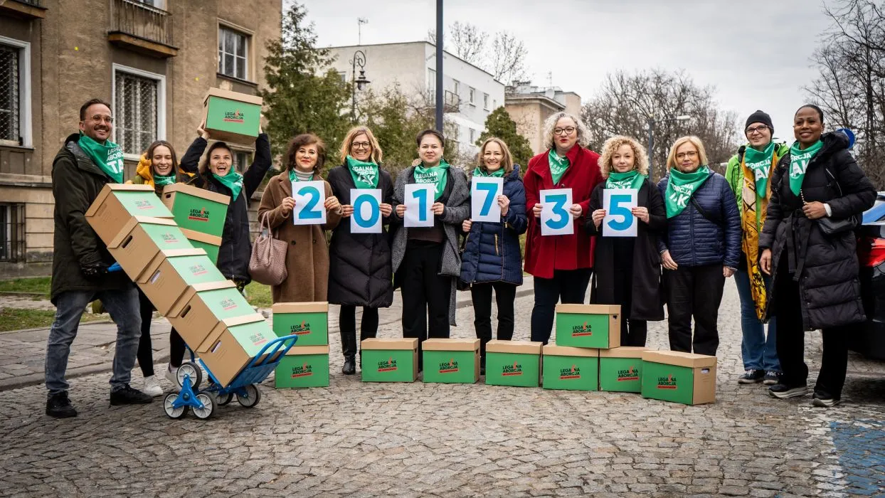 Grupa osób stojących na ulicy, trzymająca kartki z cyframi tworzącymi numer 201735 i otoczona pudełkami z napisem „LEGALNA ABORCJA”, symbolizująca zbieranie podpisów pod projektem „Legalna aborcja. Bez kompromisów”.