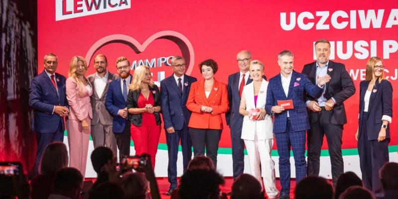 Grupa osób w eleganckich strojach stoi na scenie przy czerwonym tle z hasłem „Uczciwa praca musi popłacać” podczas wydarzenia Lewicy.