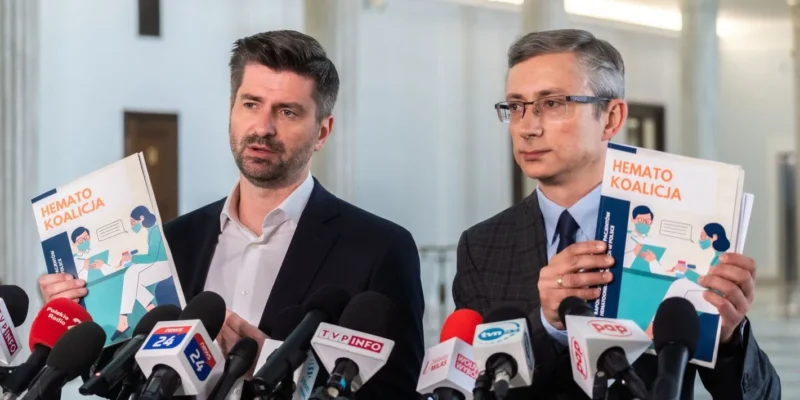 Dwóch mężczyzn w garniturach podczas konferencji prasowej trzyma ulotki z napisem „HEMATO KOALICJA” i stoi wśród mikrofonów.