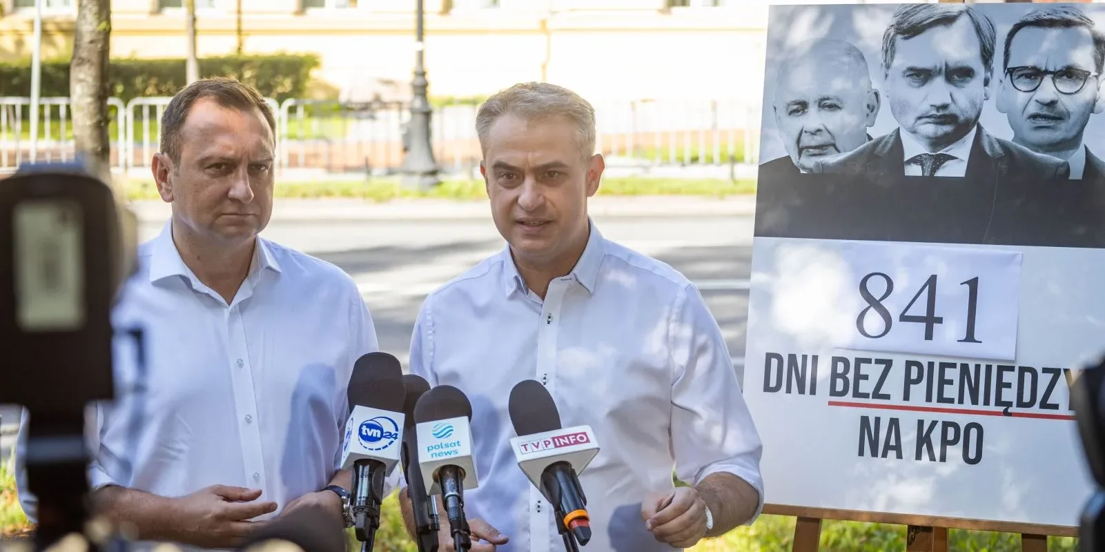 Dwóch mężczyzn w białych koszulach przemawia do mikrofonów podczas konferencji prasowej, stojąc obok plakatu z napisem „841 dni bez pieniędzy na KPO”.