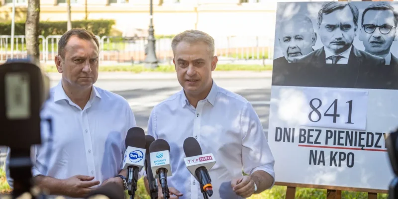 Dwóch mężczyzn w białych koszulach przemawia do mikrofonów podczas konferencji prasowej, stojąc obok plakatu z napisem „841 dni bez pieniędzy na KPO”.