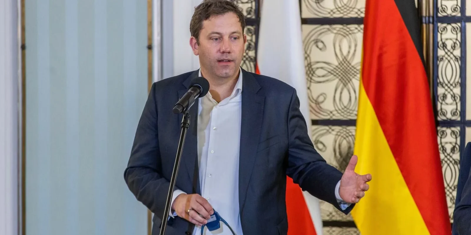 Mężczyzna w garniturze, stojący przy mikrofonie, wygłasza przemówienie na tle flag Polski i Niemiec.