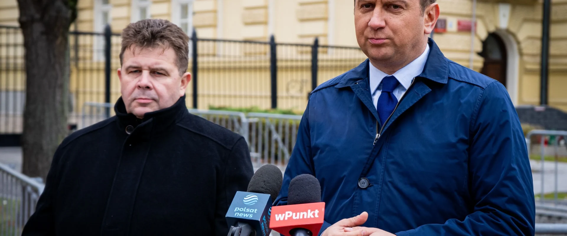 Dwóch mężczyzn stoi przy mikrofonach z logotypami Polsat News i Wpunkt podczas konferencji prasowej dotyczącej konfliktu o Turow, na tle żółtej kamienicy.