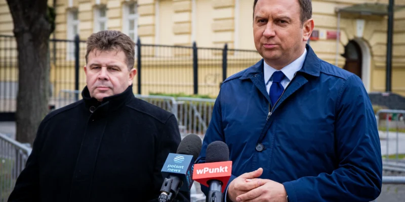 Dwóch mężczyzn stoi przy mikrofonach z logotypami Polsat News i Wpunkt podczas konferencji prasowej dotyczącej konfliktu o Turow, na tle żółtej kamienicy.