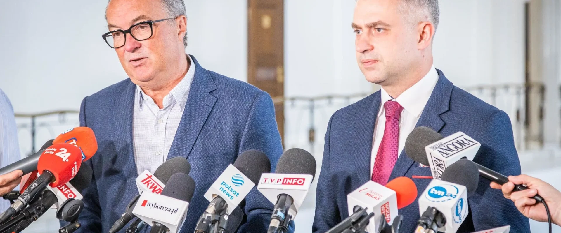 Dwaj mężczyźni w garniturach stoją przed mikrofonami podczas konferencji prasowej w jasnym wnętrzu.