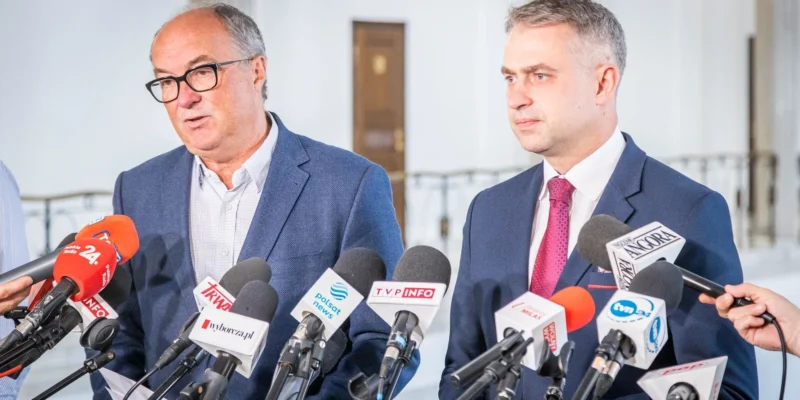 Dwaj mężczyźni w garniturach stoją przed mikrofonami podczas konferencji prasowej w jasnym wnętrzu.