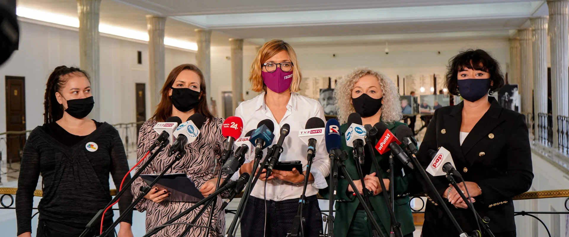 Na zdjęciu widać grupę kobiet stojących przed mikrofonami podczas konferencji prasowej w przestronnym wnętrzu.