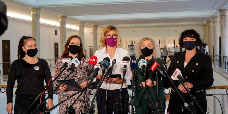 Na zdjęciu widać grupę kobiet stojących przed mikrofonami podczas konferencji prasowej w przestronnym wnętrzu.