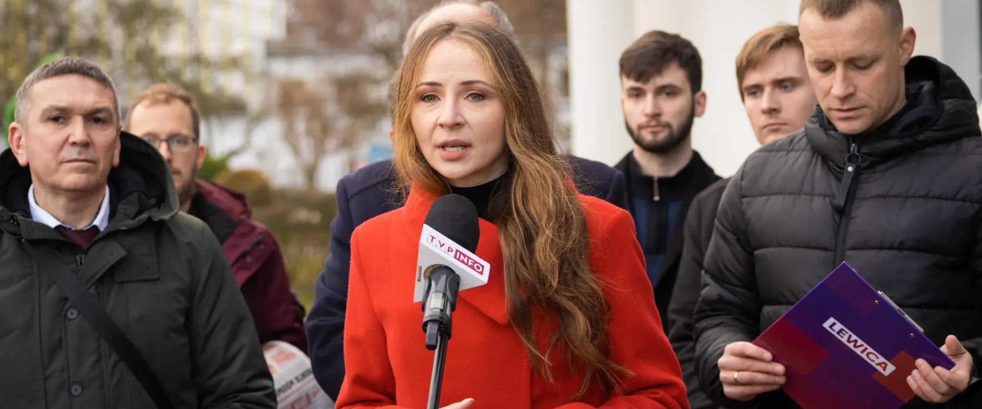 Na zdjęciu widoczna jest kobieta w czerwonym płaszczu, mówiąca do mikrofonu, otoczona przez grupę mężczyzn, z tłem w postaci budynków i drzew.
