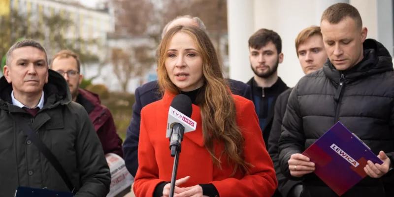 Na zdjęciu widoczna jest kobieta w czerwonym płaszczu, mówiąca do mikrofonu, otoczona przez grupę mężczyzn, z tłem w postaci budynków i drzew.
