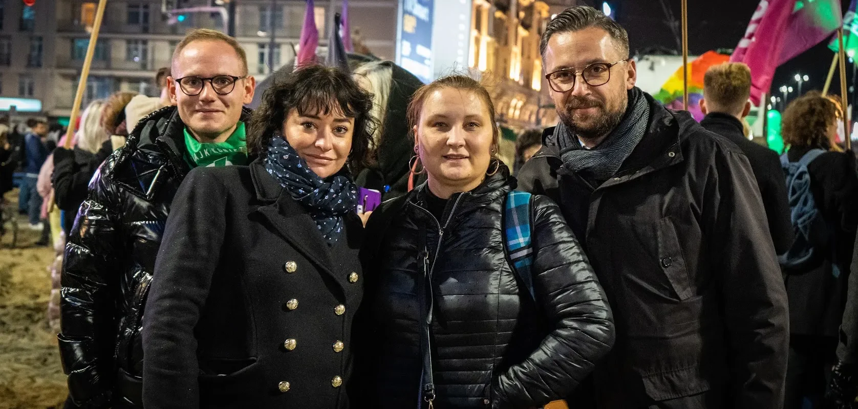 Grupa czterech osób pozująca do zdjęcia na tle wieczornego miasta, podczas wydarzenia publicznego.