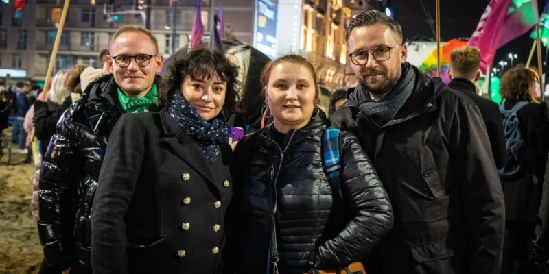 Grupa czterech osób pozująca do zdjęcia na tle wieczornego miasta, podczas wydarzenia publicznego.