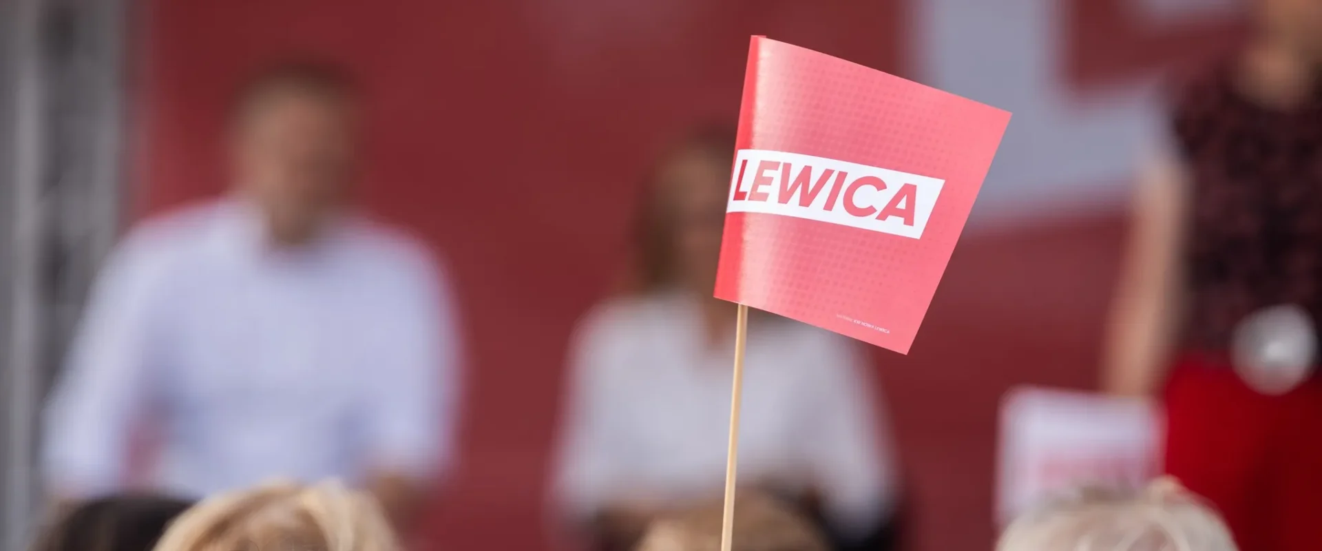 Czerwona kartka z białym napisem LEWICA na patyku widoczna wśród rozmytego tłumu podczas inauguracji kampanii Lewicy.