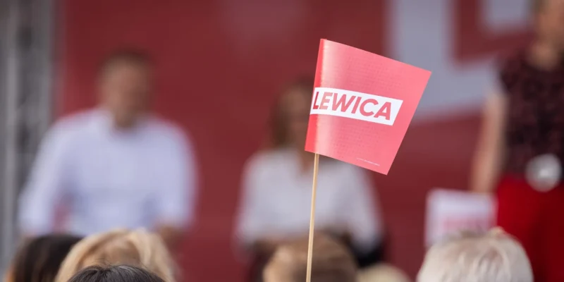 Czerwona kartka z białym napisem LEWICA na patyku widoczna wśród rozmytego tłumu podczas inauguracji kampanii Lewicy.