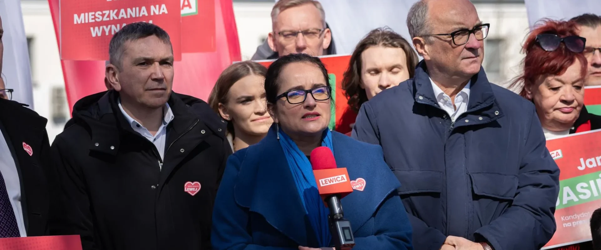 Grupa ludzi na wiecu politycznym, kobieta w niebieskim płaszczu przemawia do mikrofonu z logiem Lewicy, otoczona czerwonymi plakatami z hasłami o mieszkaniach na wynajem.