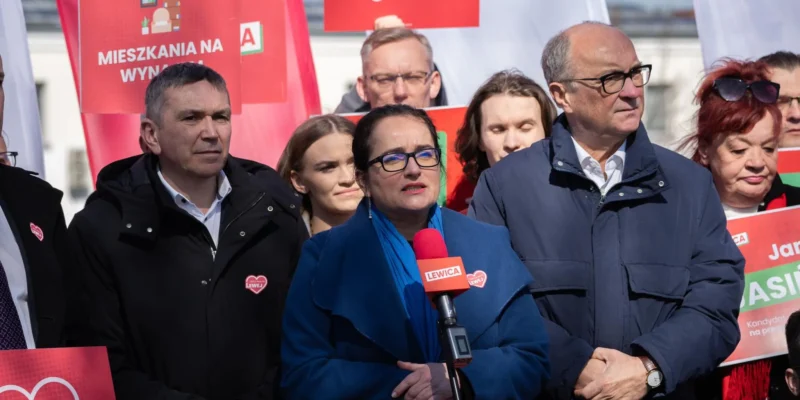 Grupa ludzi na wiecu politycznym, kobieta w niebieskim płaszczu przemawia do mikrofonu z logiem Lewicy, otoczona czerwonymi plakatami z hasłami o mieszkaniach na wynajem.