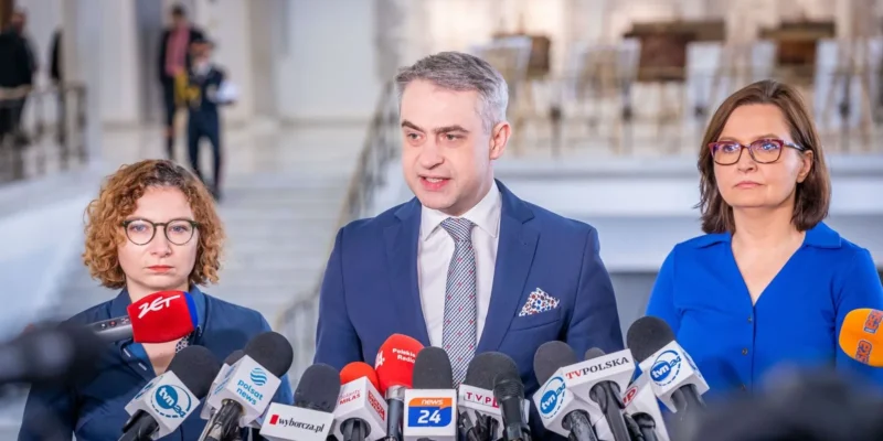 Trójka osób w garniturach i strojach formalnych stoi za mikrofonami prasowymi podczas konferencji, w tle widać schody i półmrok wnętrza budynku.