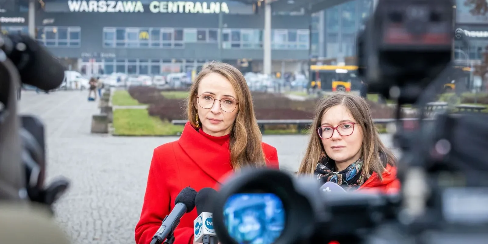 Dwie kobiety w czerwonych kurtkach stoją przed kamerami i mikrofonami, udzielając konferencji prasowej na placu przed dworcem Warszawa Centralna w sprawie zamrożenia cen biletów kolejowych.