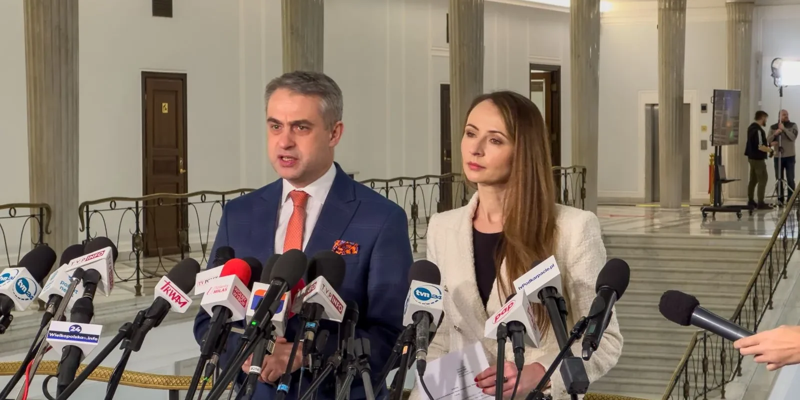 Na zdjęciu widać dwóch mówców, mężczyznę i kobietę, stojących przed mikrofonami podczas konferencji prasowej w eleganckim wnętrzu.