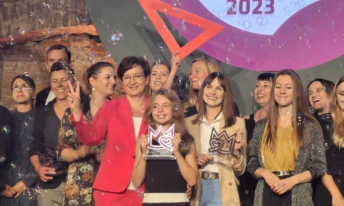 Grupa osób na scenie świętująca wręczenie nagrody Korony Równości, a na pierwszym planie kobieta w różowym garniturze trzyma trofeum, wokół latają bańki mydlane i w tle widać baner z napisem Równość 2023.