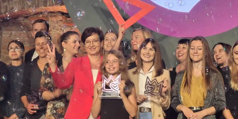 Grupa osób na scenie świętująca wręczenie nagrody Korony Równości, a na pierwszym planie kobieta w różowym garniturze trzyma trofeum, wokół latają bańki mydlane i w tle widać baner z napisem Równość 2023.