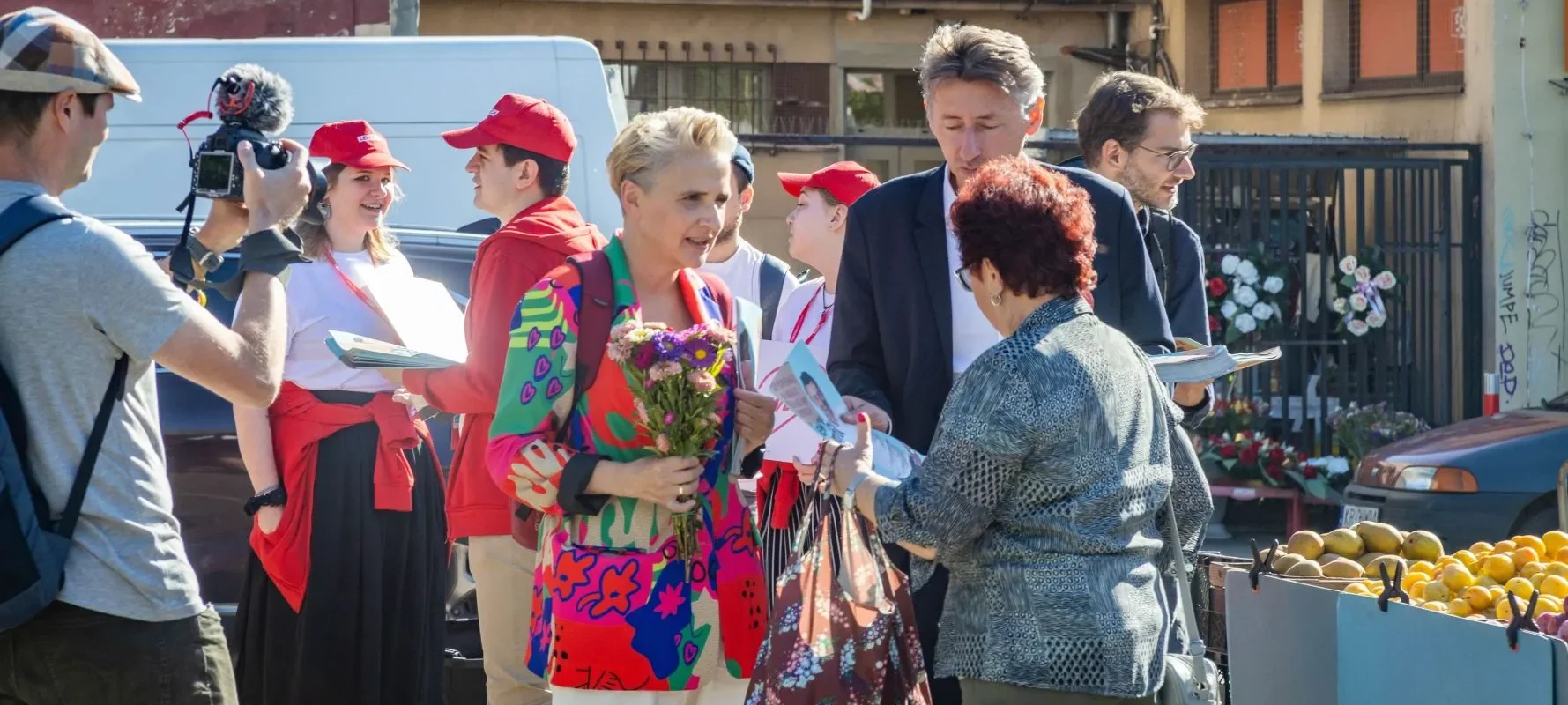 Grupa ludzi na targowisku, wśród nich kobieta trzymająca bukiet kwiatów i dwie osoby w czerwonych czapkach, rozmawia z dziennikarzem z kamerą podczas wydarzenia prasowego Lewicy dotyczącego ustawy o zawodzie artysty.