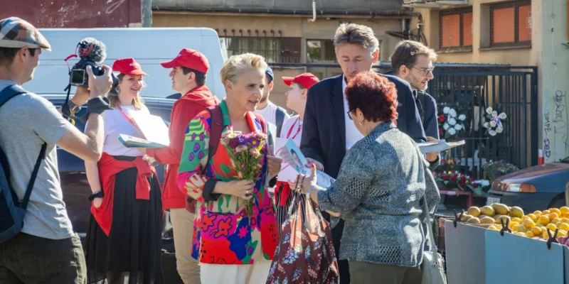 Grupa ludzi na targowisku, wśród nich kobieta trzymająca bukiet kwiatów i dwie osoby w czerwonych czapkach, rozmawia z dziennikarzem z kamerą podczas wydarzenia prasowego Lewicy dotyczącego ustawy o zawodzie artysty.