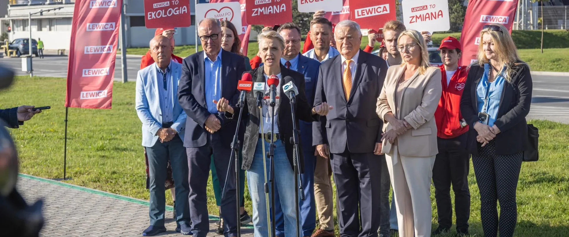 Kobieta przemawiająca do mikrofonów podczas konferencji prasowej Lewicy na zewnątrz, wśród grupy działaczy i czerwonych flag z napisem LEWICA.