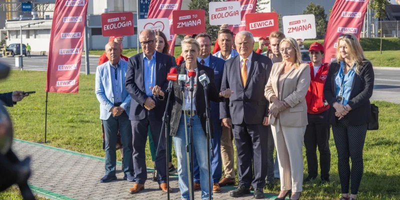 Kobieta przemawiająca do mikrofonów podczas konferencji prasowej Lewicy na zewnątrz, wśród grupy działaczy i czerwonych flag z napisem LEWICA.