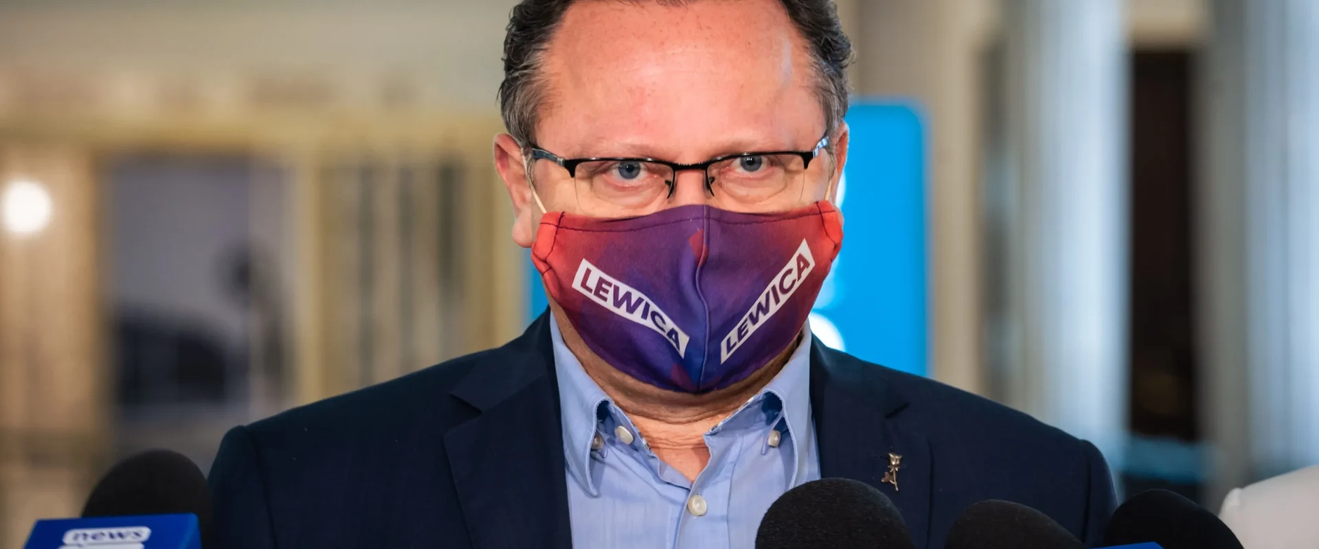 Mężczyzna w garniturze i okularach, noszący maseczkę z napisem LEWICA, przemawia do mediów podczas konferencji prasowej.
