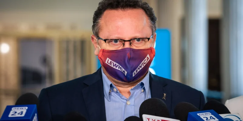 Mężczyzna w garniturze i okularach, noszący maseczkę z napisem LEWICA, przemawia do mediów podczas konferencji prasowej.