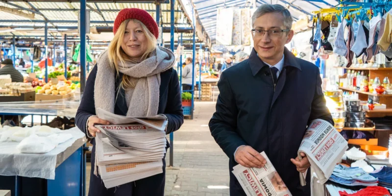 Dwoje dorosłych na targowisku, kobieta w czerwonej czapce i szalu trzyma stos gazet, a mężczyzna w okularach również trzyma gazetę, w tle stragany z warzywami, odzieżą i innymi artykułami.