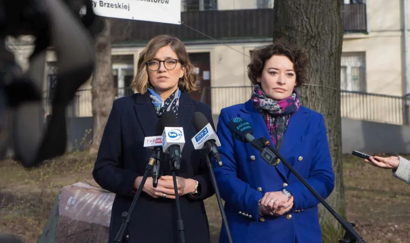 Dwie kobiety stojące przed mikrofonami podczas konferencji prasowej na zewnątrz, jedna w granatowym płaszczu z okularami, druga w niebieskim płaszczu z kolorowym szalem, w tle drzewa i budynki.