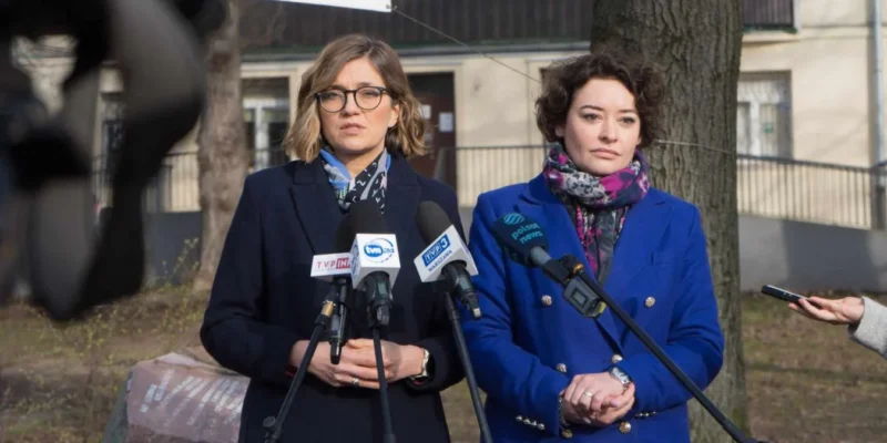 Dwie kobiety stojące przed mikrofonami podczas konferencji prasowej na zewnątrz, jedna w granatowym płaszczu z okularami, druga w niebieskim płaszczu z kolorowym szalem, w tle drzewa i budynki.