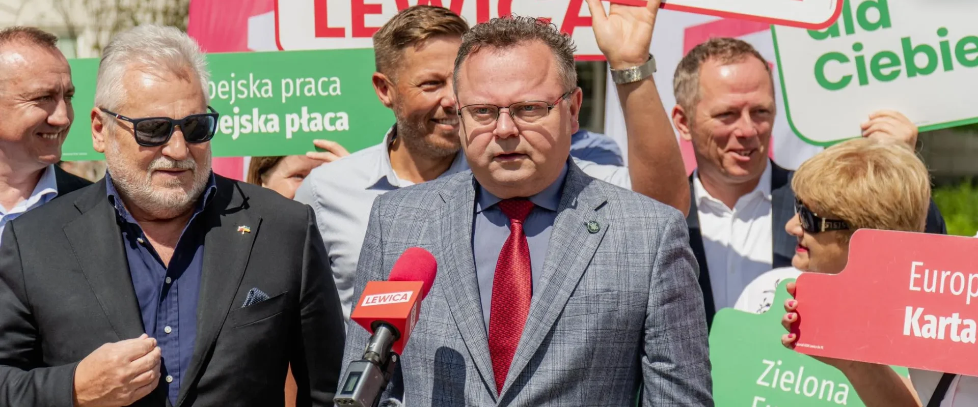 Mężczyzna w szaro-niebieskim garniturze i czerwonym krawacie przemawia do mikrofonu podczas konferencji Lewicy, w otoczeniu plakatów z hasłami o Silnej Europie i współpracy w zakresie bezpieczeństwa.