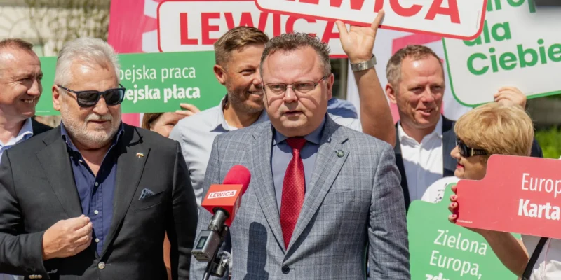 Mężczyzna w szaro-niebieskim garniturze i czerwonym krawacie przemawia do mikrofonu podczas konferencji Lewicy, w otoczeniu plakatów z hasłami o Silnej Europie i współpracy w zakresie bezpieczeństwa.