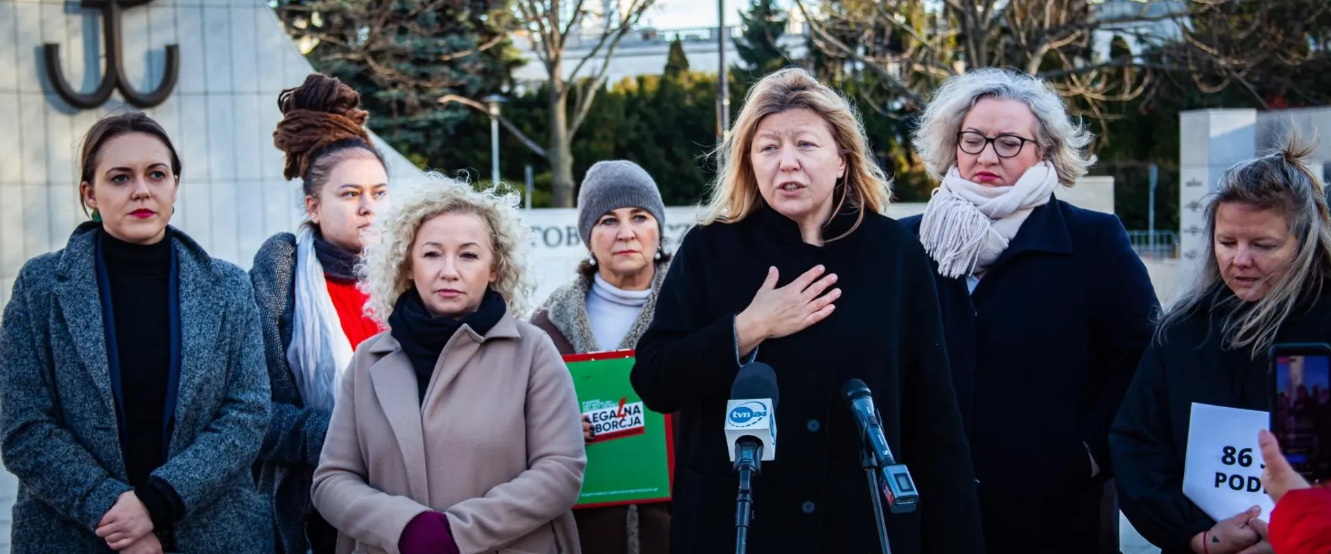 Kobiety w różnorodnych strojach stoją przy mównicy, niektóre z nich trzymają dokumenty, tło przedstawia symbol związany z historią Polski.