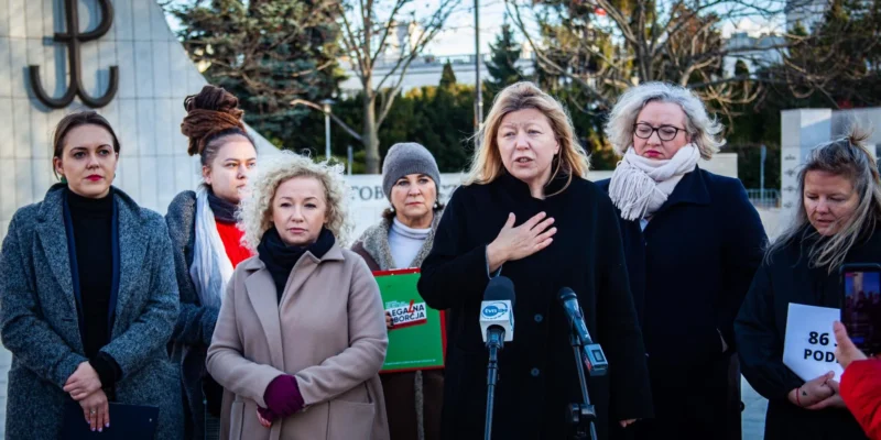 Kobiety w różnorodnych strojach stoją przy mównicy, niektóre z nich trzymają dokumenty, tło przedstawia symbol związany z historią Polski.