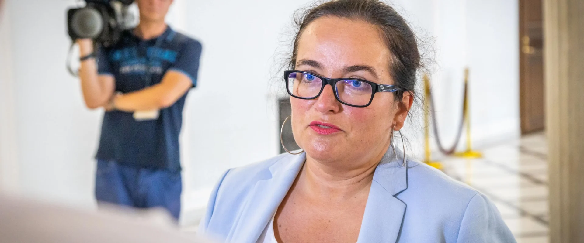 Kobieta w jasnoniebieskim garniturze i czarnych okularach udziela wywiadu; w tle stoi operator z kamerą.