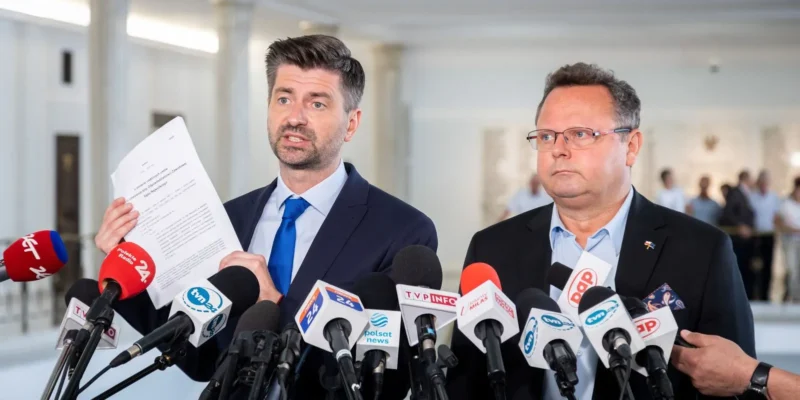 Na zdjęciu dwóch mężczyzn stoi przed mikrofonami, jeden z nich trzyma dokument i prowadzi konferencję prasową.