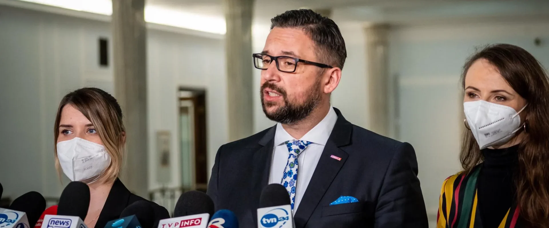 Mężczyzna w garniturze i okularach przemawia do mediów podczas konferencji prasowej, otoczony przez dwie kobiety w maseczkach.